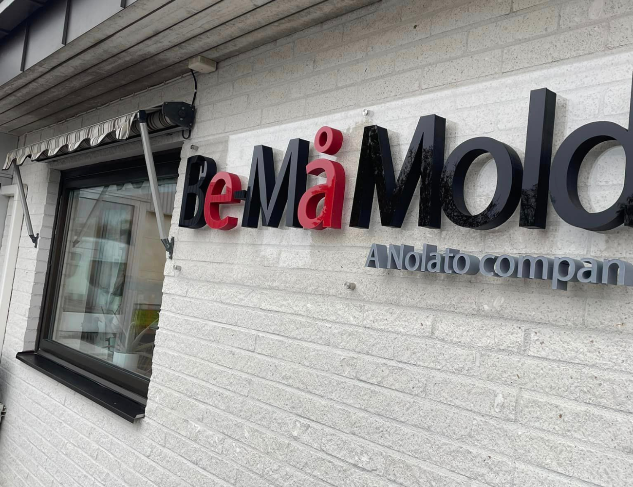En svart och röd skylt med följande text "BE-MÅ MOLD" som sitter på en husfasad. Grå text under "A nalato Company"